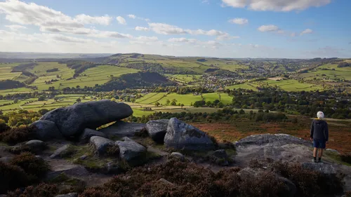The landscape photograph captures Curbar Edge View in Derbyshire, United Kingdom, during the early afternoon in autumn. The scene showcases the rugged natural beauty of the Peak District, with expansive rural fields, rolling hills, and patchwork farmland stretching into the distance. Large rock formations are prominent in the foreground, characteristic of Curbar Edge, while the lush countryside is dotted with scattered trees and hedgerows. A person stands near the edge, observing the landscape, which highlights the harmonious relationship between humans and nature in this part of the United Kingdom. The photograph provides a clear illustration of the rural scenery for which Derbyshire and the Peak District are renowned.